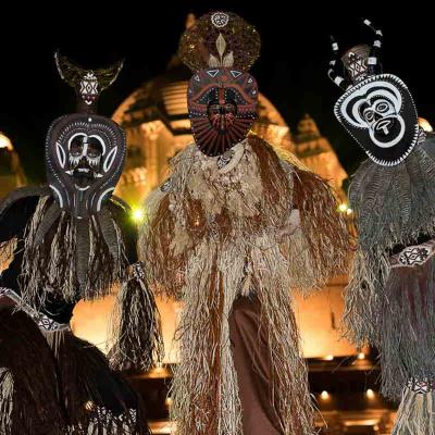 Three performers wearing traditional-style masks and straw costumes standing outdoors at night in front of an illuminated building.