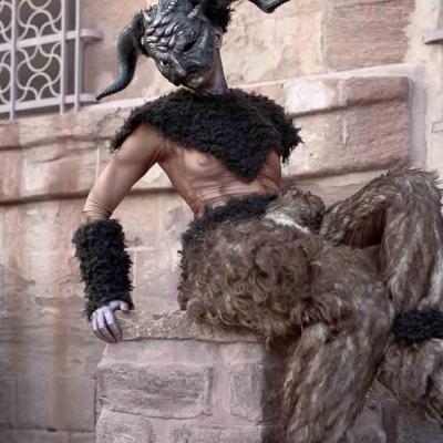 Person wearing a horned mask and furry costume, sitting on stone steps in front of a stone building.