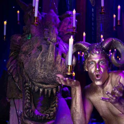 Performer with gold body paint and ram horns posing beside a large dragon head under hanging candles.