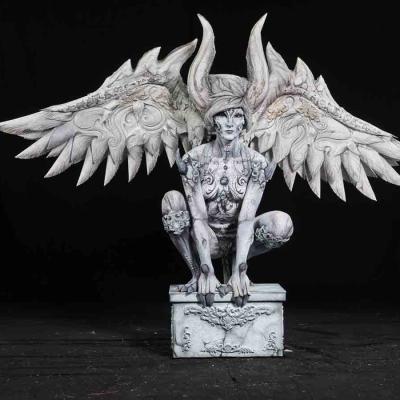 Person painted like a stone statue with large wings and horns, crouching on a pedestal against a black background.